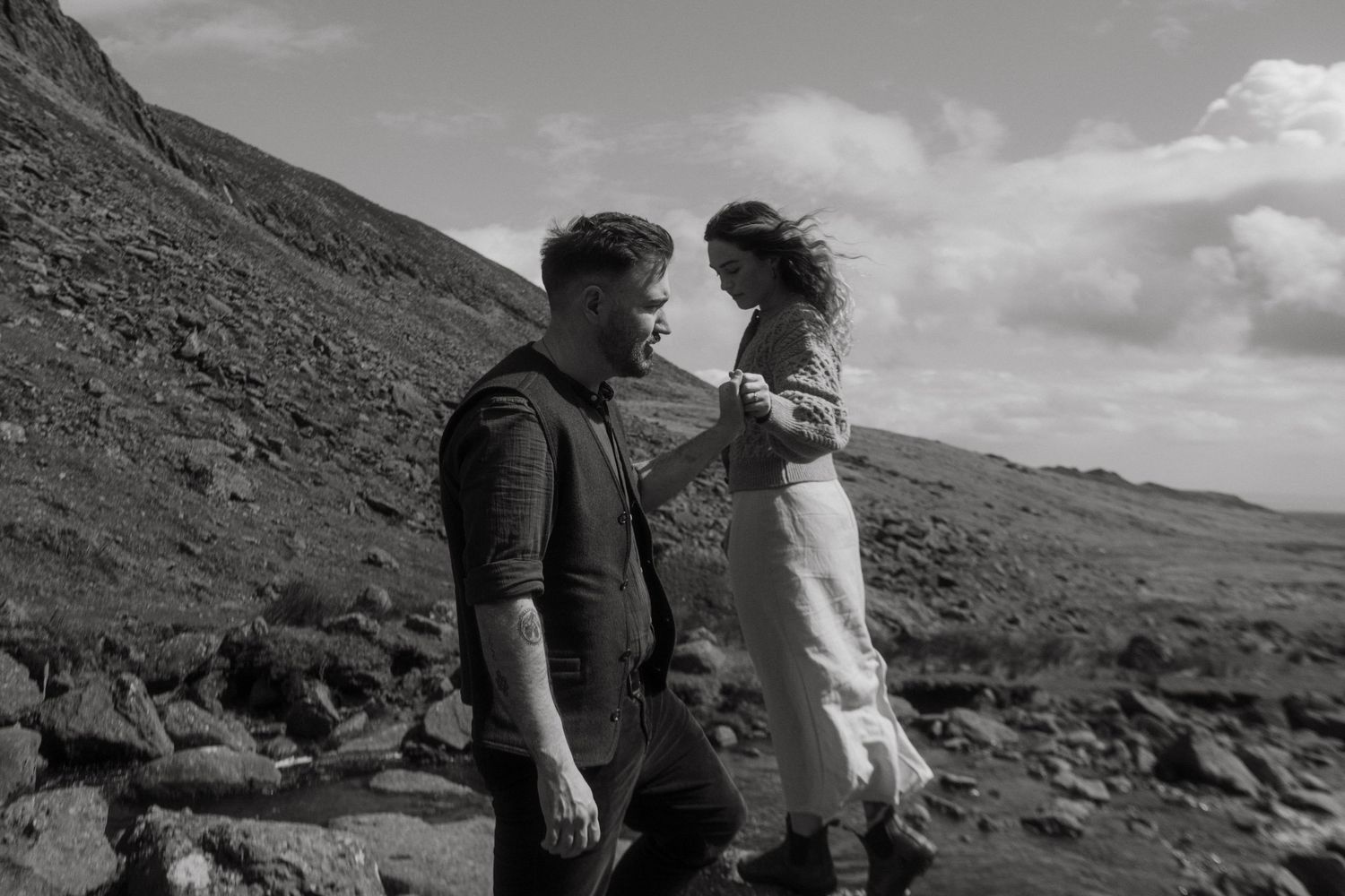 Black and white photo of a couple, he's holding her hand while balancing barefoot on rocks
