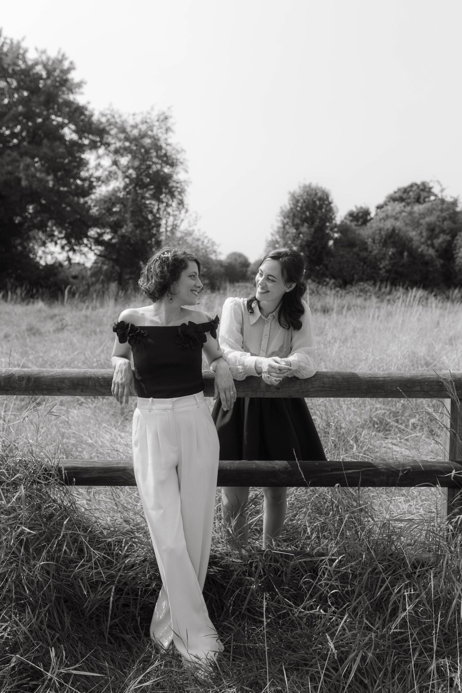 Black and white photograph of two brides looking at each other while leaning on a fence after their civic ceremony