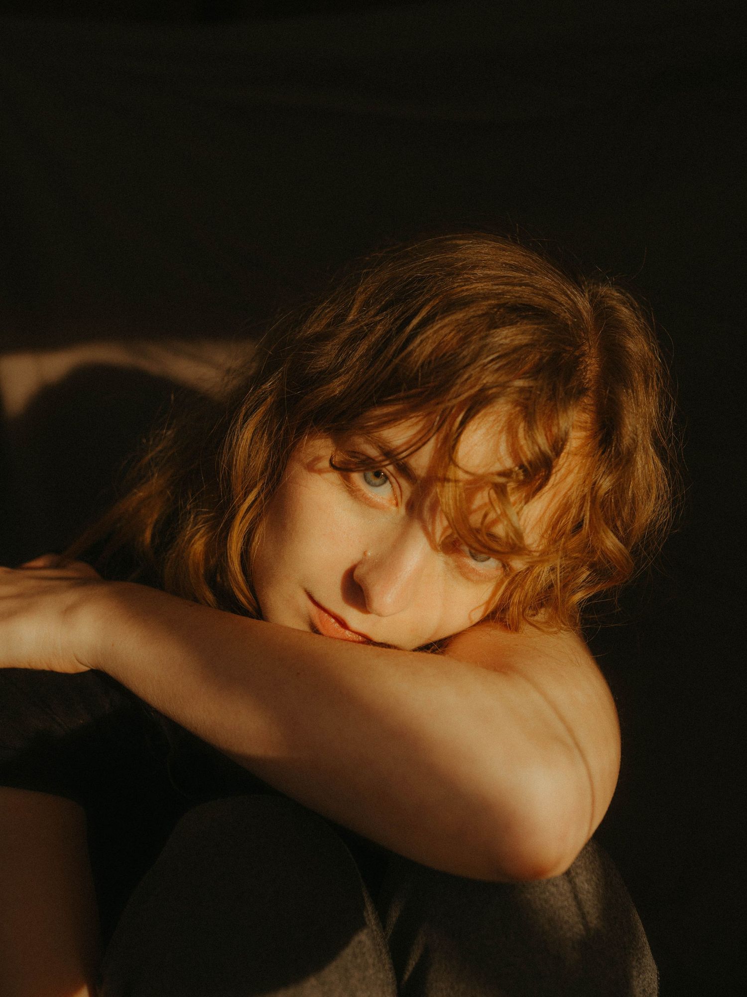 Self Portrait of the photographer in direct sunlight, leaning with her arm against her leg. Blonde curly hair, black background