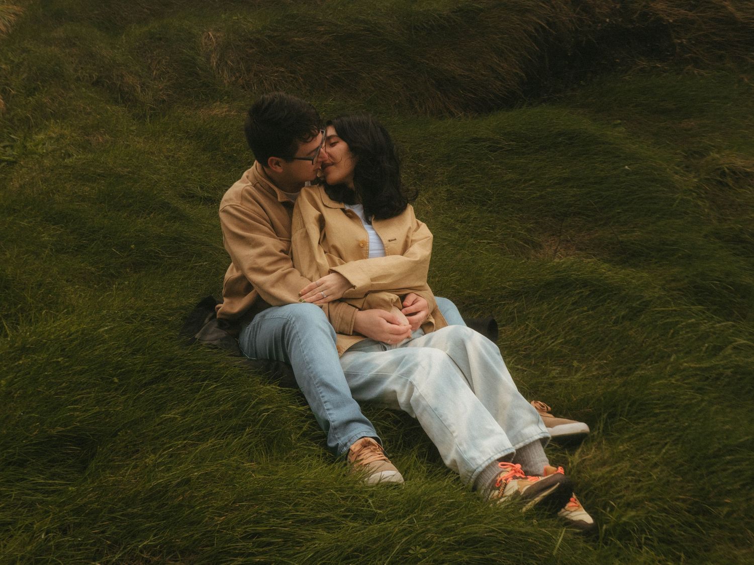 Photograph of couple sitting in a meadow of tall grass, she's sitting between his legs and they are kissing