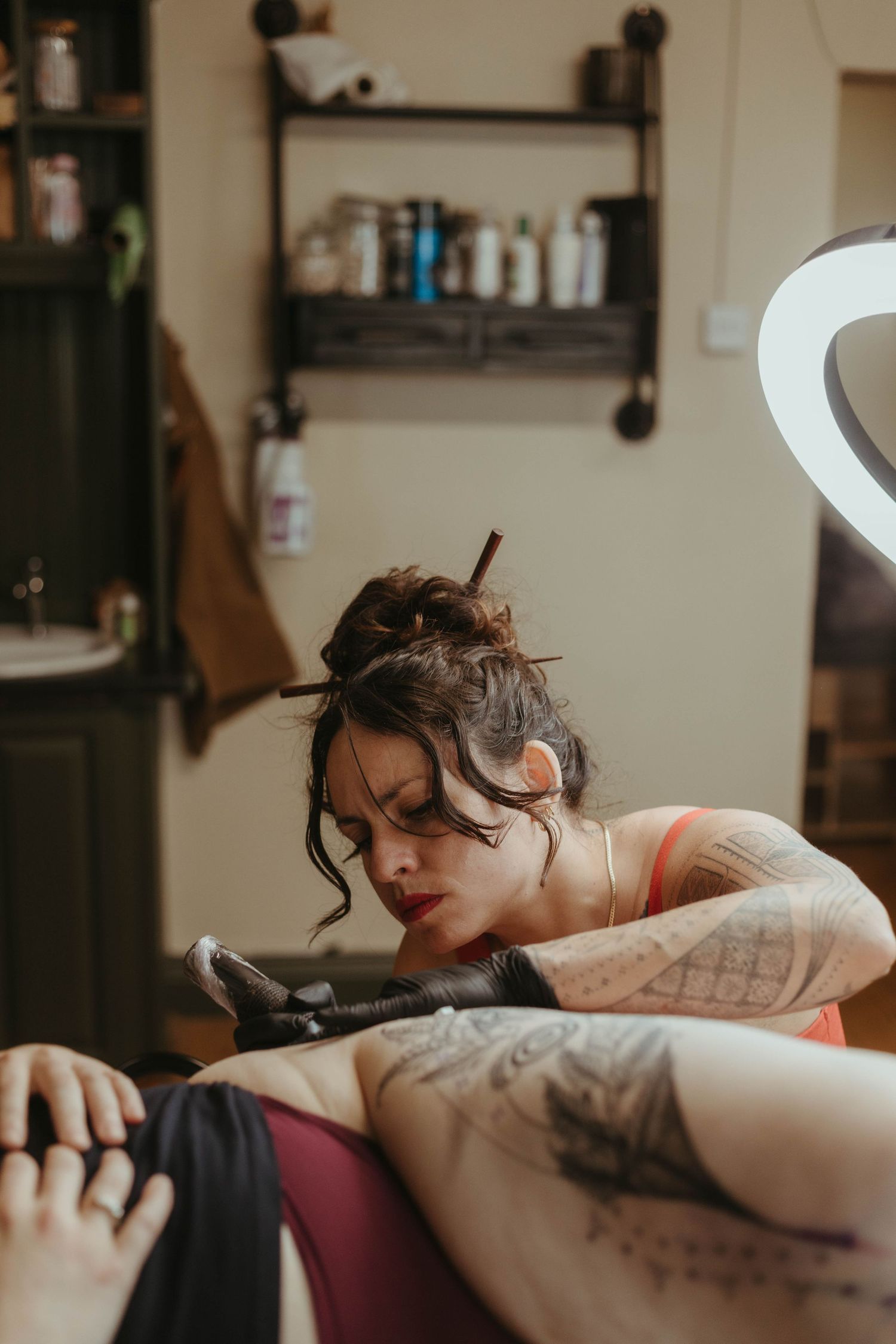 Photo of tattoo artist tattooing a client, she has her hair tied up and is wearing a red top