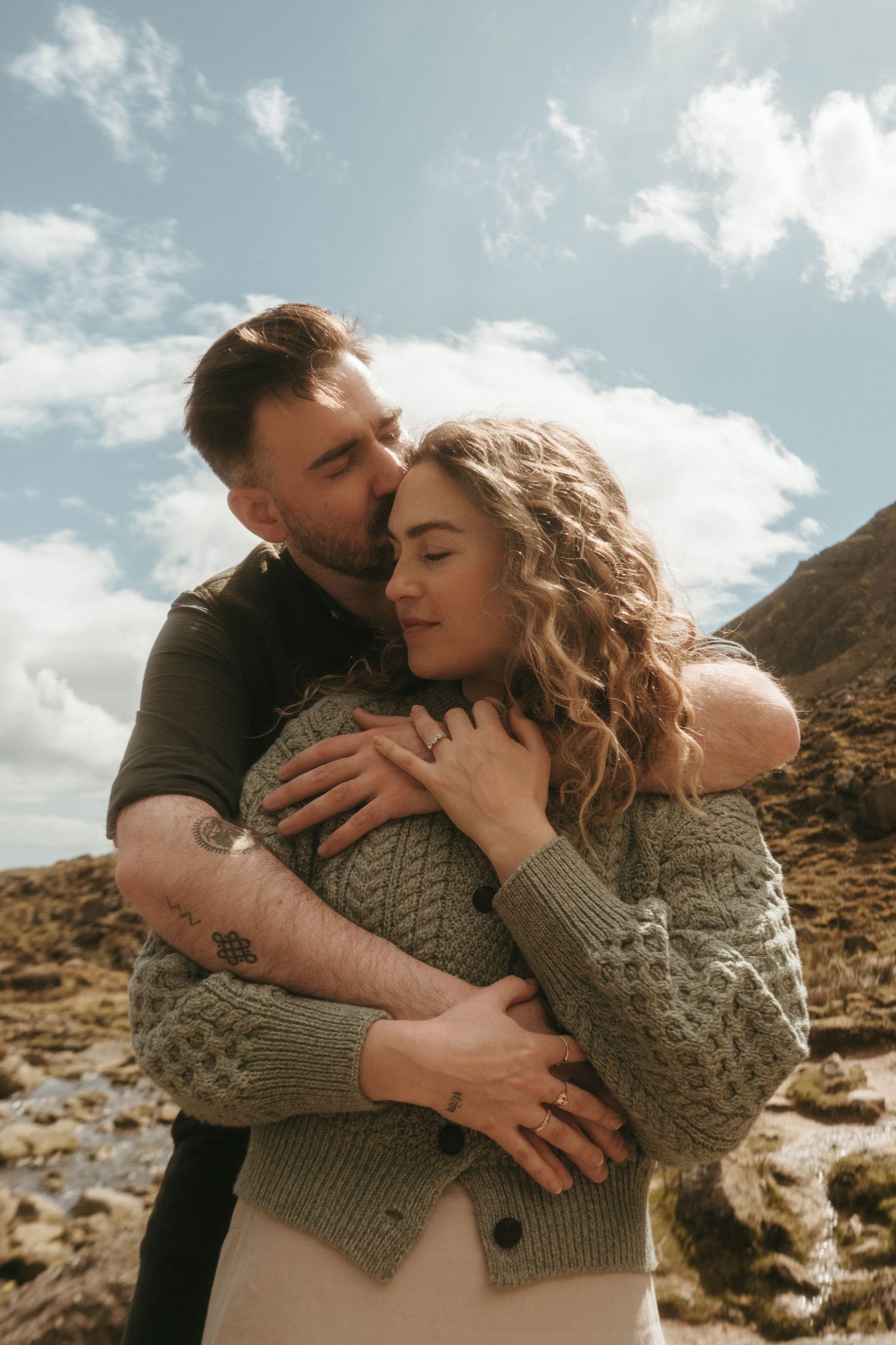 picture of couple in front of rugged green mountain, she's leaning against him and his eyes are closed