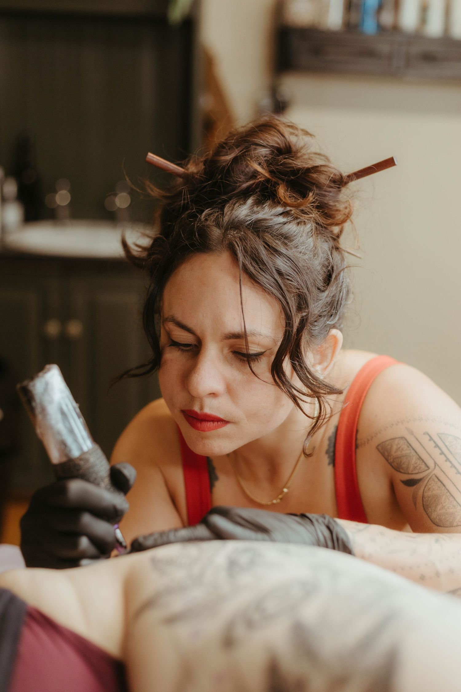 Photo of tattoo artist tattooing a client, she has her hair tied up and is wearing a red top