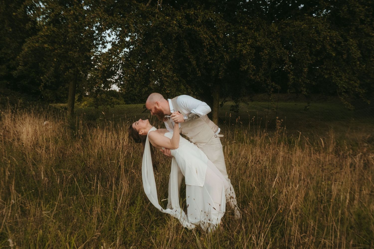 Photograph of bride and groom in a meadow after their wedding ceremony, she has dark brown hair and he is bald with a ginger beard.