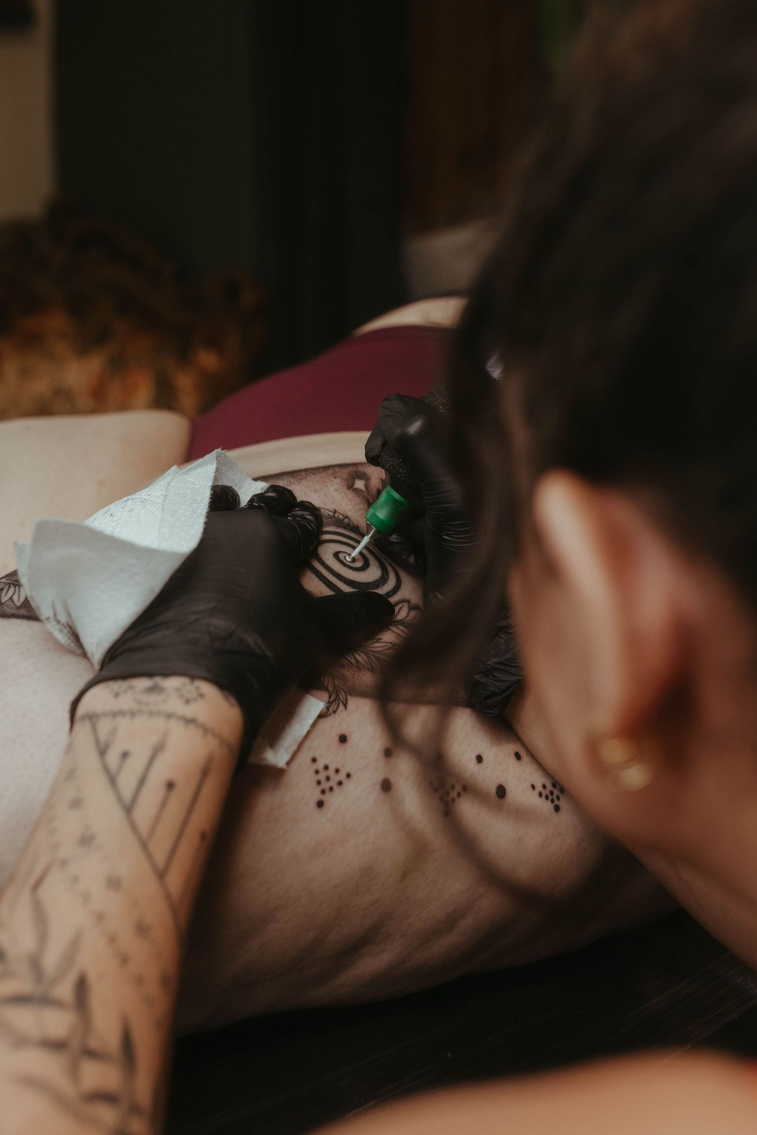 Photo of tattoo artist tattooing a client, she has her hair tied up and is wearing a red top