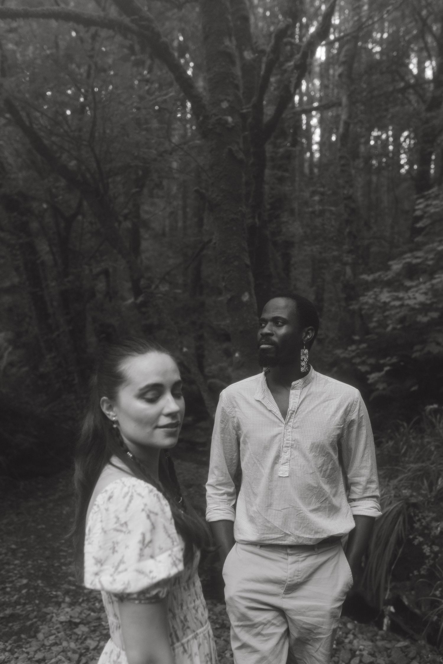 Black and white photograph of a couple in a forest very soft shadows, he's standing behind her
