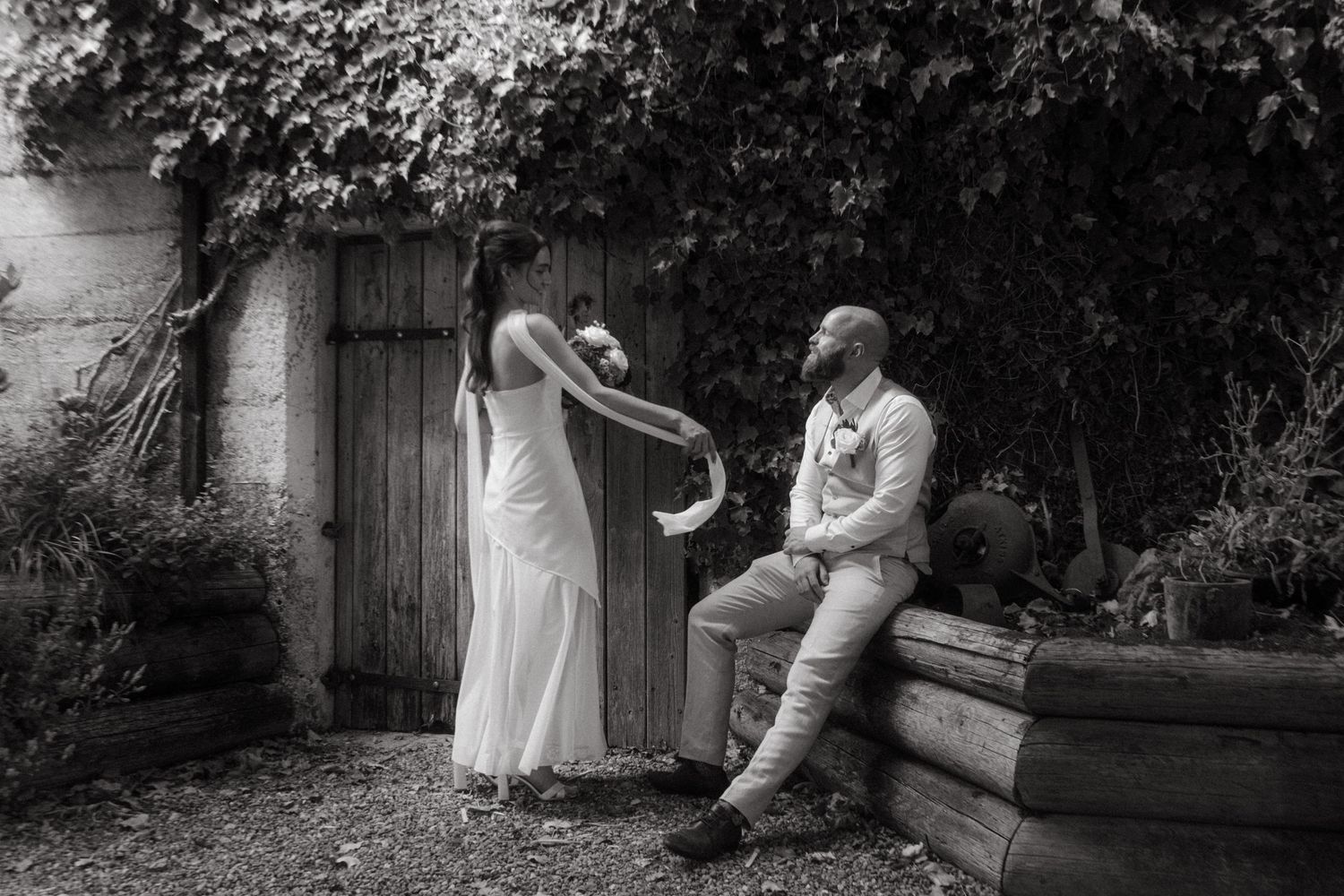 Black and white photograph of bride and groom after their weddin, he's sitting against logs watching her spin in her dress.