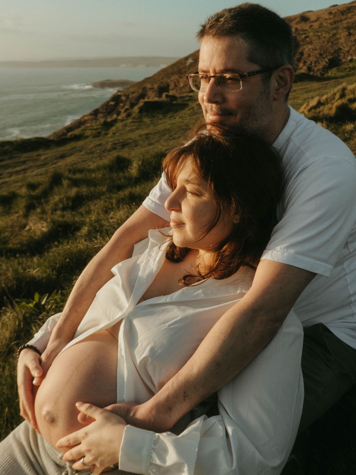 Photograph of a couple on a cliff outlooking the sea, she's leaning against him, they are sitting