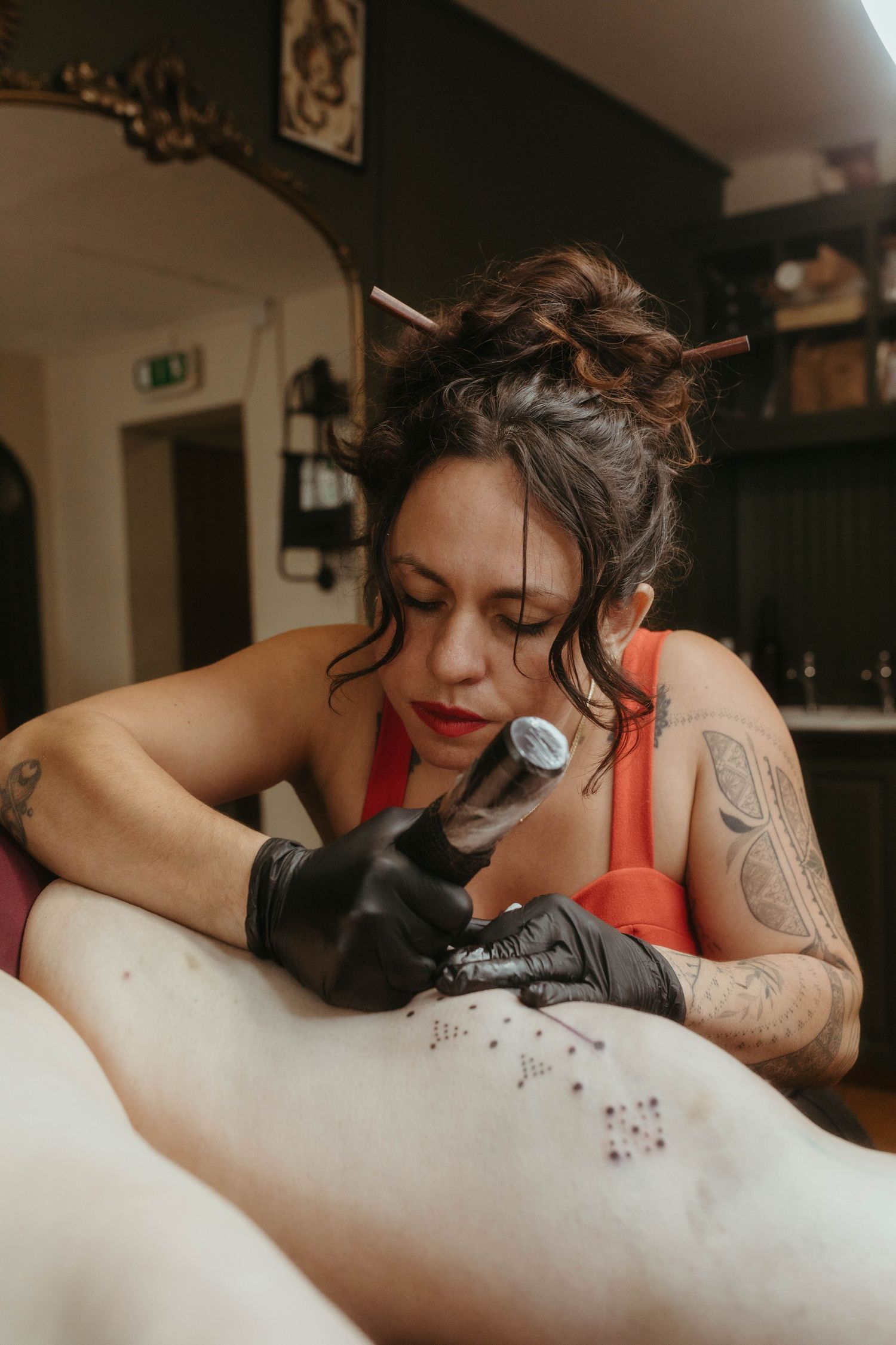 Photo of tattoo artist tattooing a client, she has her hair tied up and is wearing a red top