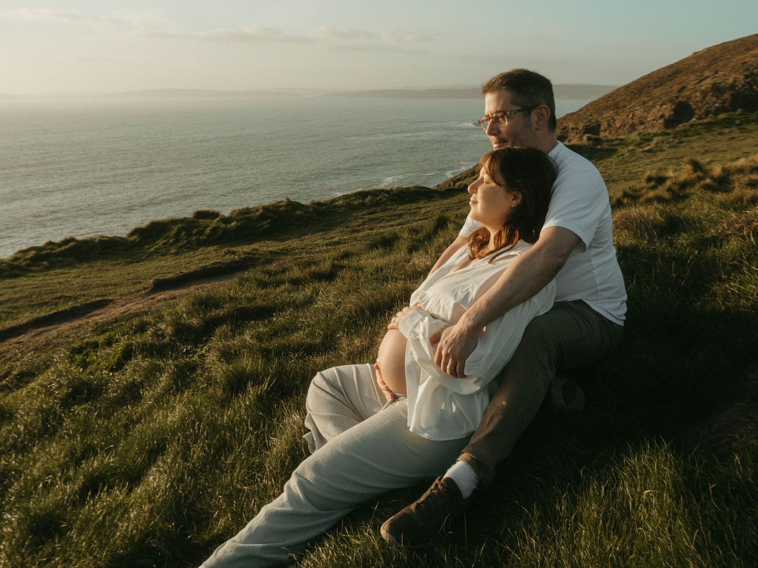 Photograph of a couple on a cliff outlooking the sea, she's leaning against him, they are sitting