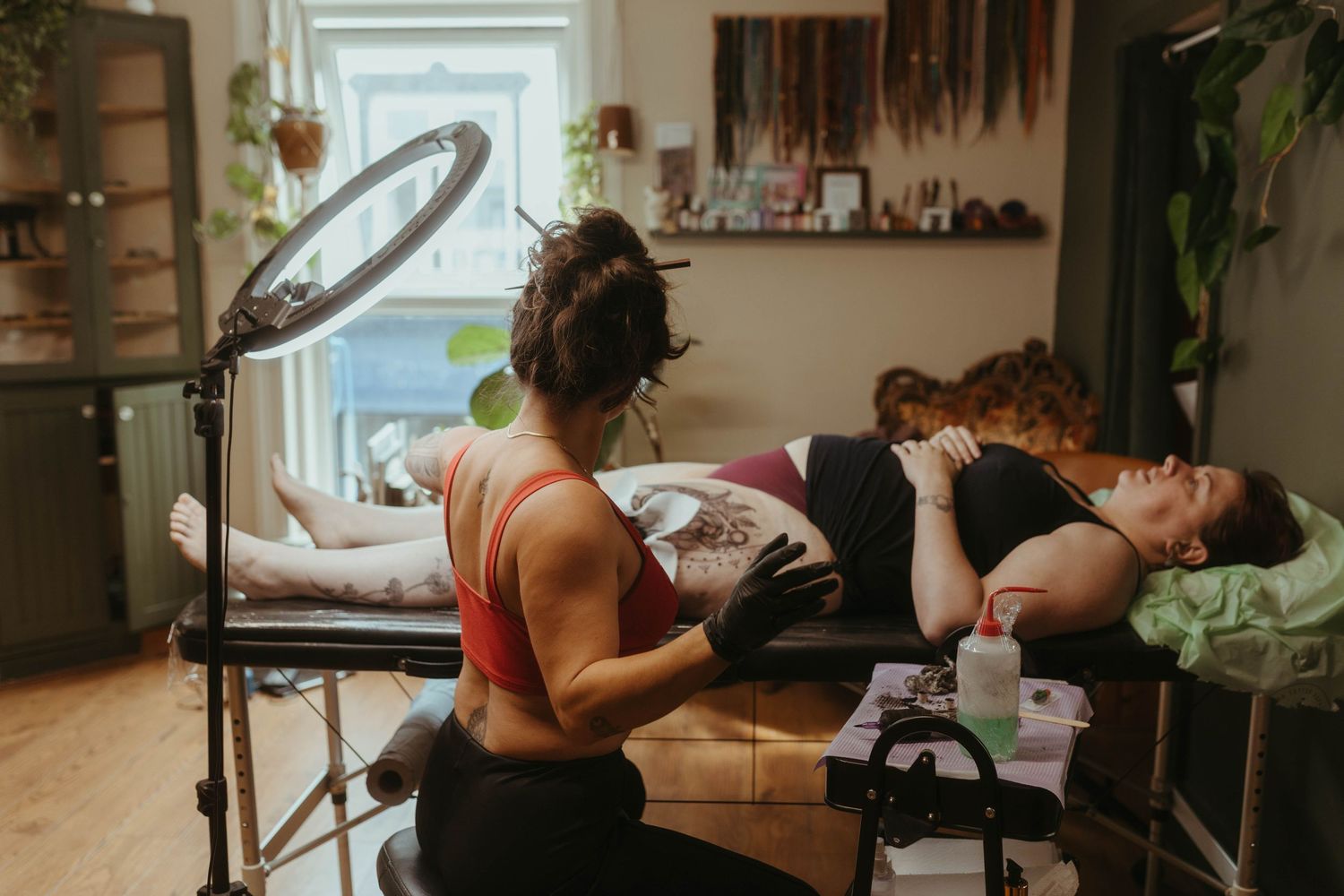 Photo of tattoo artist tattooing a client, she has her hair tied up and is wearing a red top