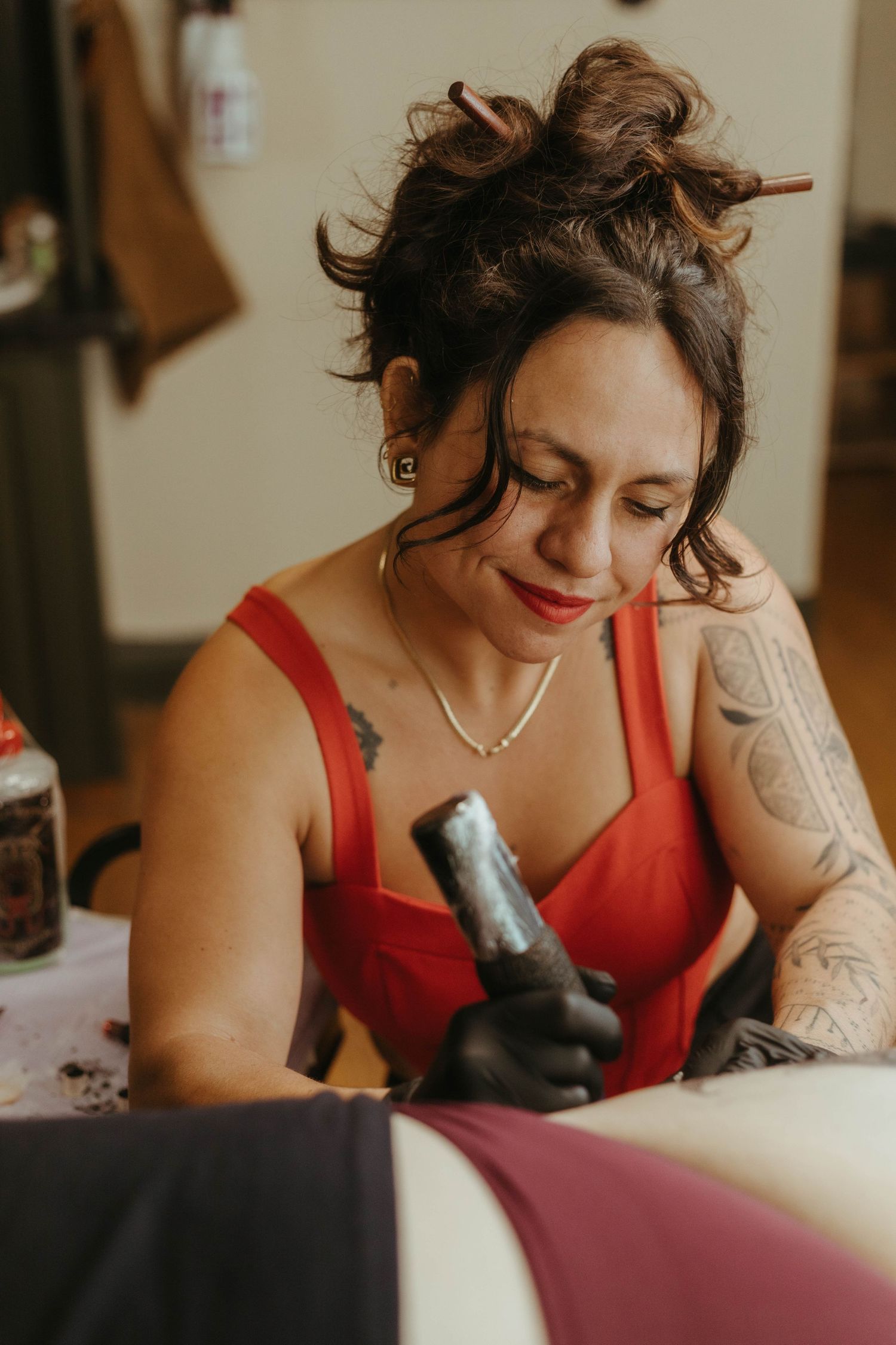 Photo of tattoo artist tattooing a client, she has her hair tied up and is wearing a red top