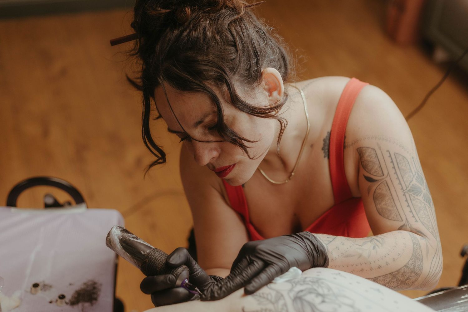 Photo of tattoo artist tattooing a client, she has her hair tied up and is wearing a red top