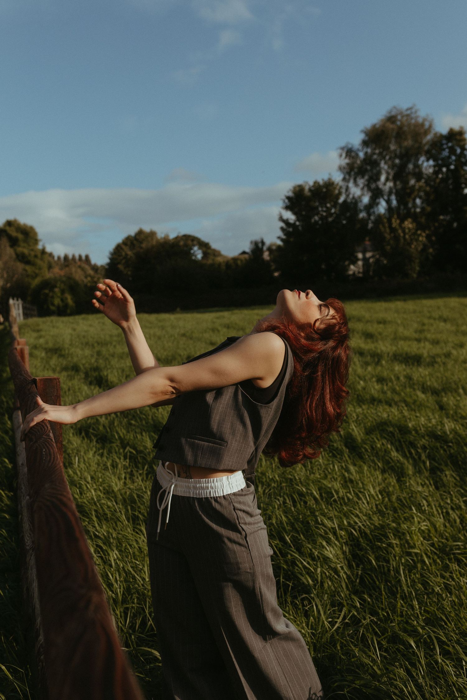 Color Photograph of a model with red flowy hair, she is leaning back while holding on to a fence, in front of a green Irish field
