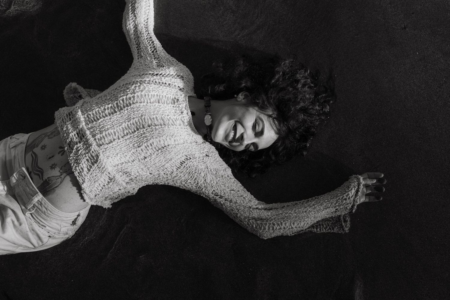 Black and white top down shot of a model laying in the sand, you can see her stomach tattoo.