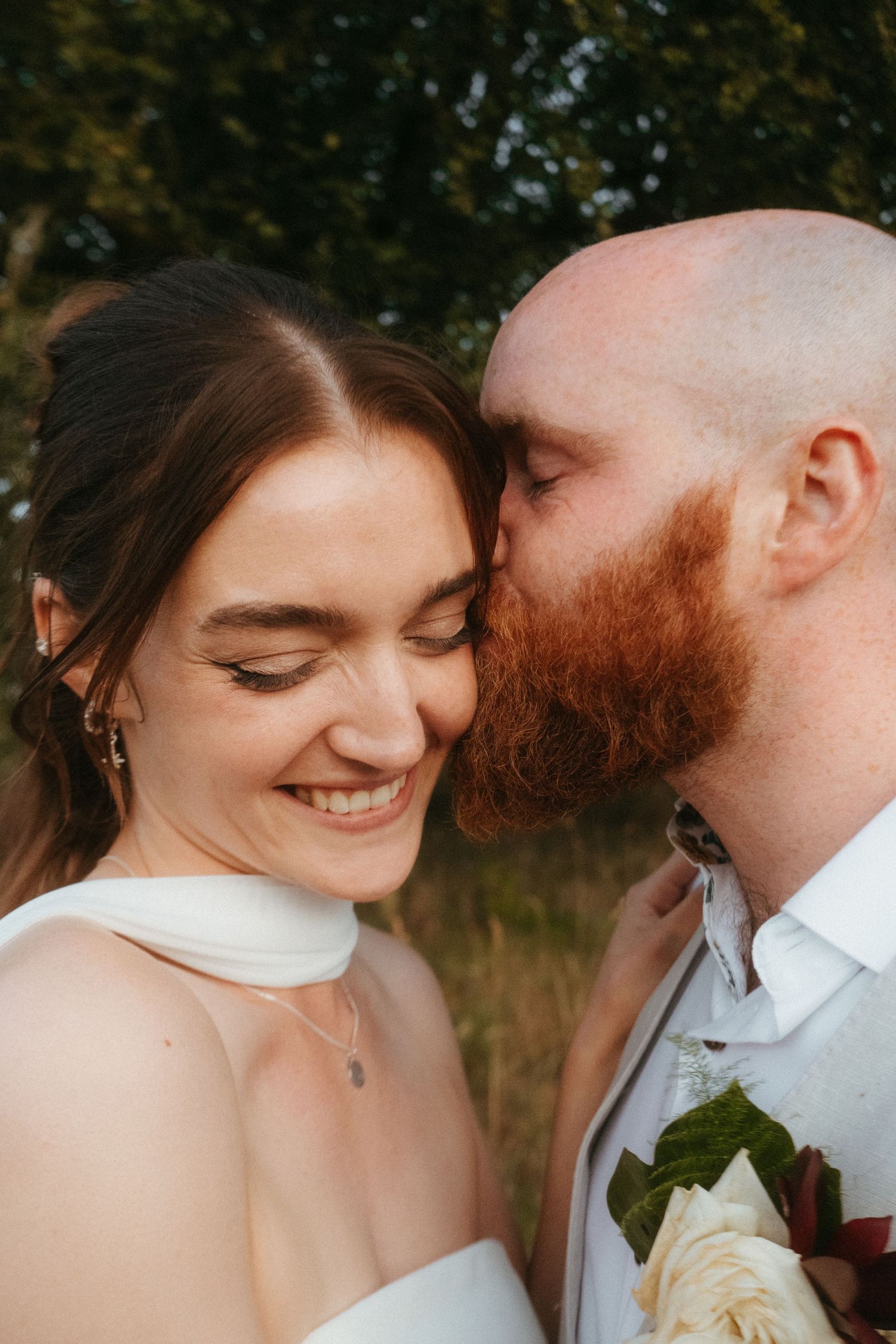 Photograph of bride and groom in a meadow after their wedding ceremony, she has dark brown hair and he is bald with a ginger beard.