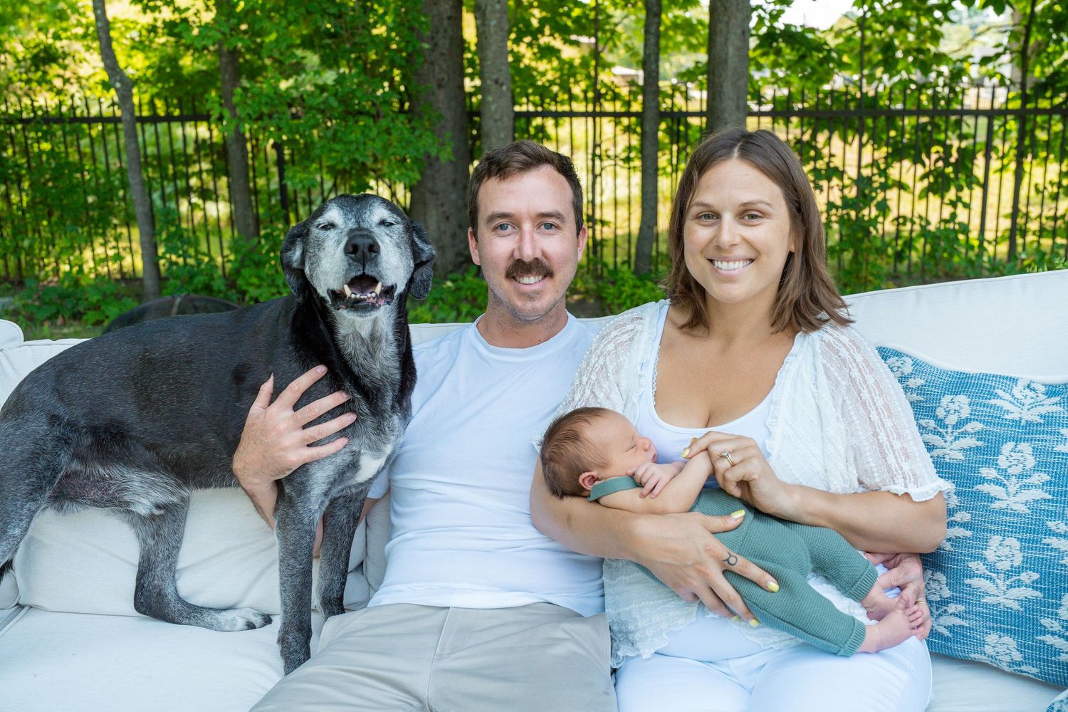 A couple with a newborn and their senior dog pose together on an outdoor patio sofa.