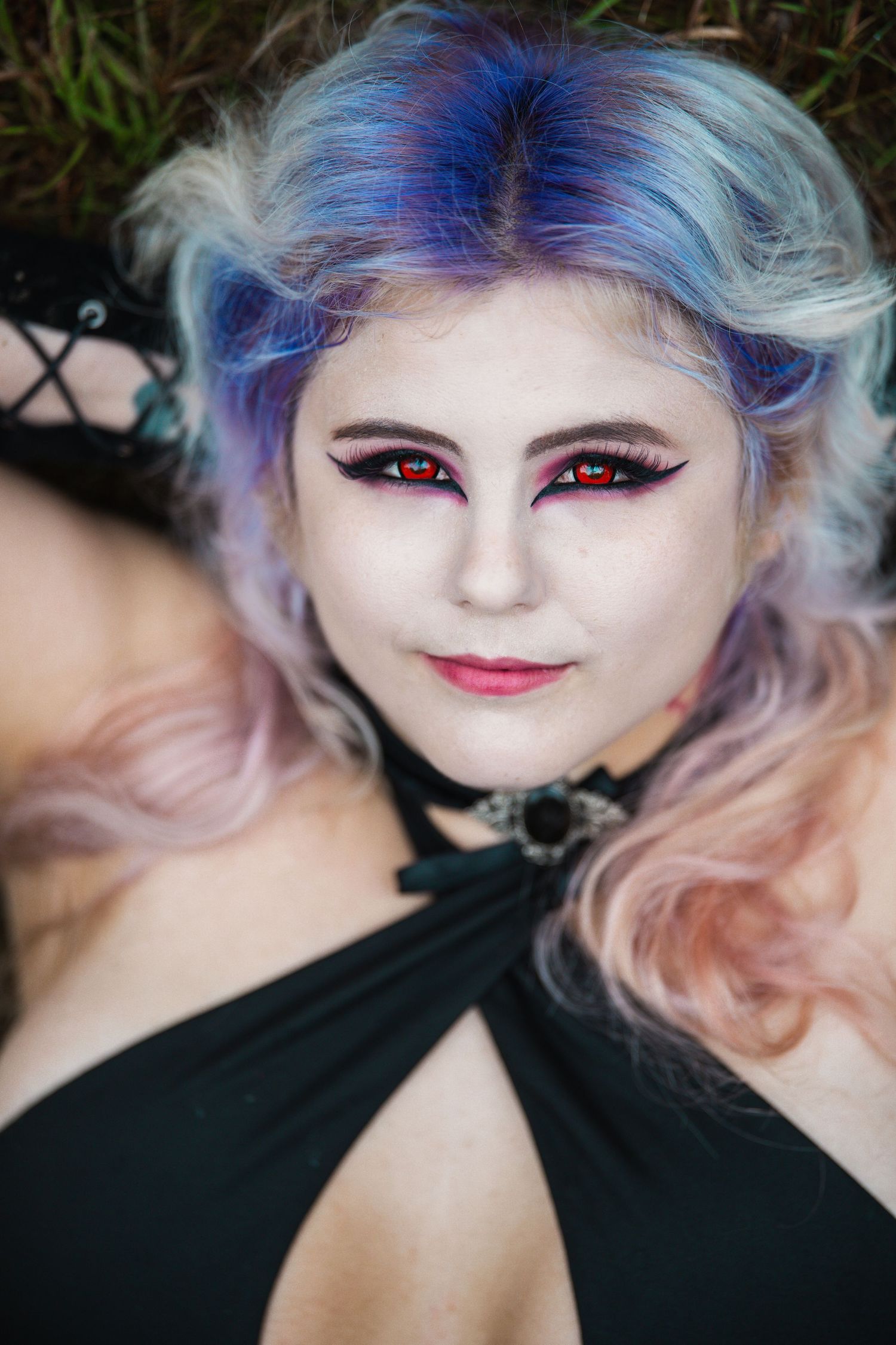 Woman with colorful blue-pink hair, red contact lenses, dramatic gothic makeup, and black choker lying in grass.