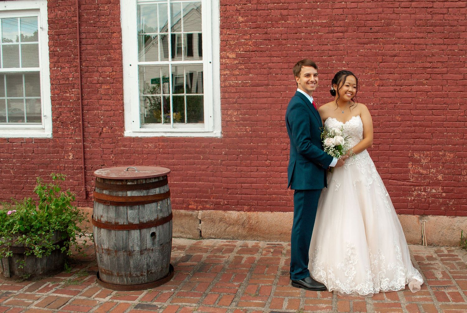 Bride and groom smiling together outside a red brick building with a rustic wooden barrel nearby.