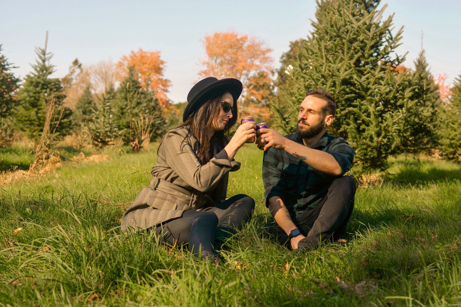 Couple sitting on grass toasting drinks at a Christmas tree farm in autumn sunlight.