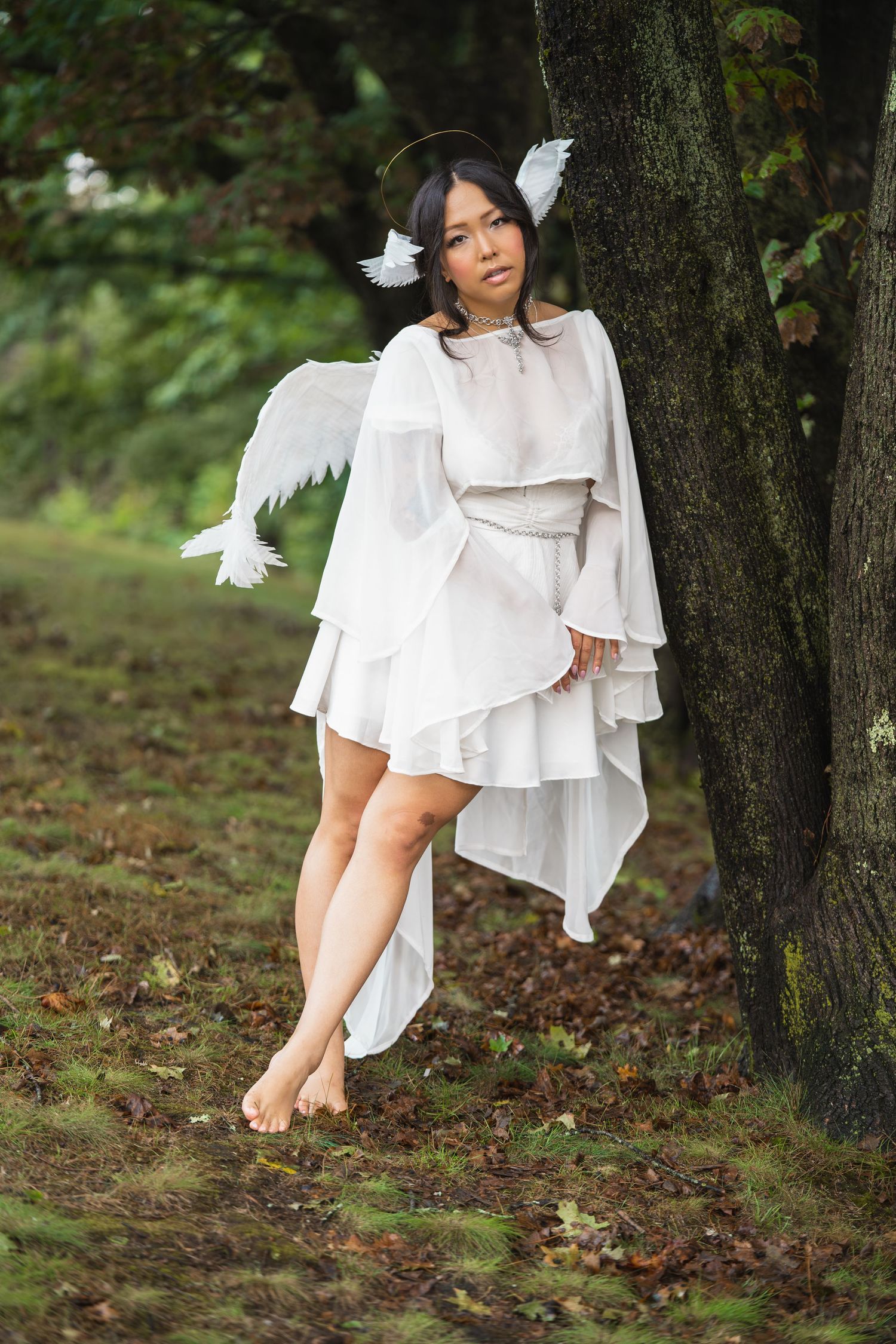 Young woman in white angel costume with wings and halo poses barefoot against a tree in a forest setting.