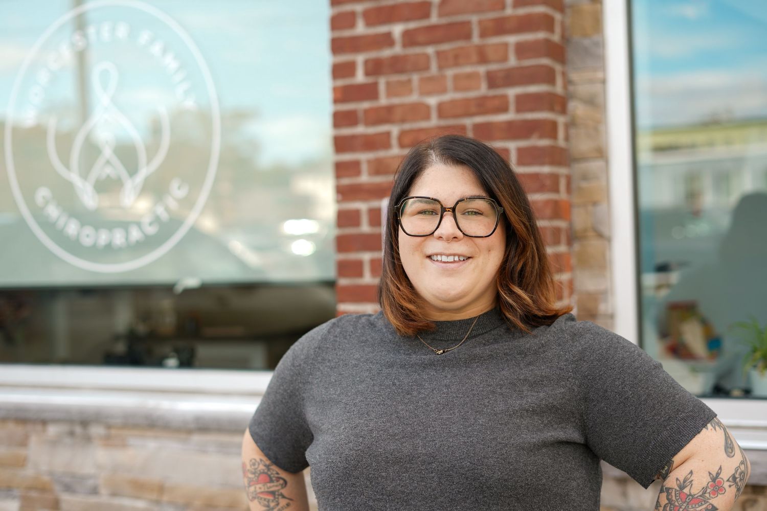 Smiling woman with glasses and tattoos stands outside a chiropractic office in a gray t-shirt.