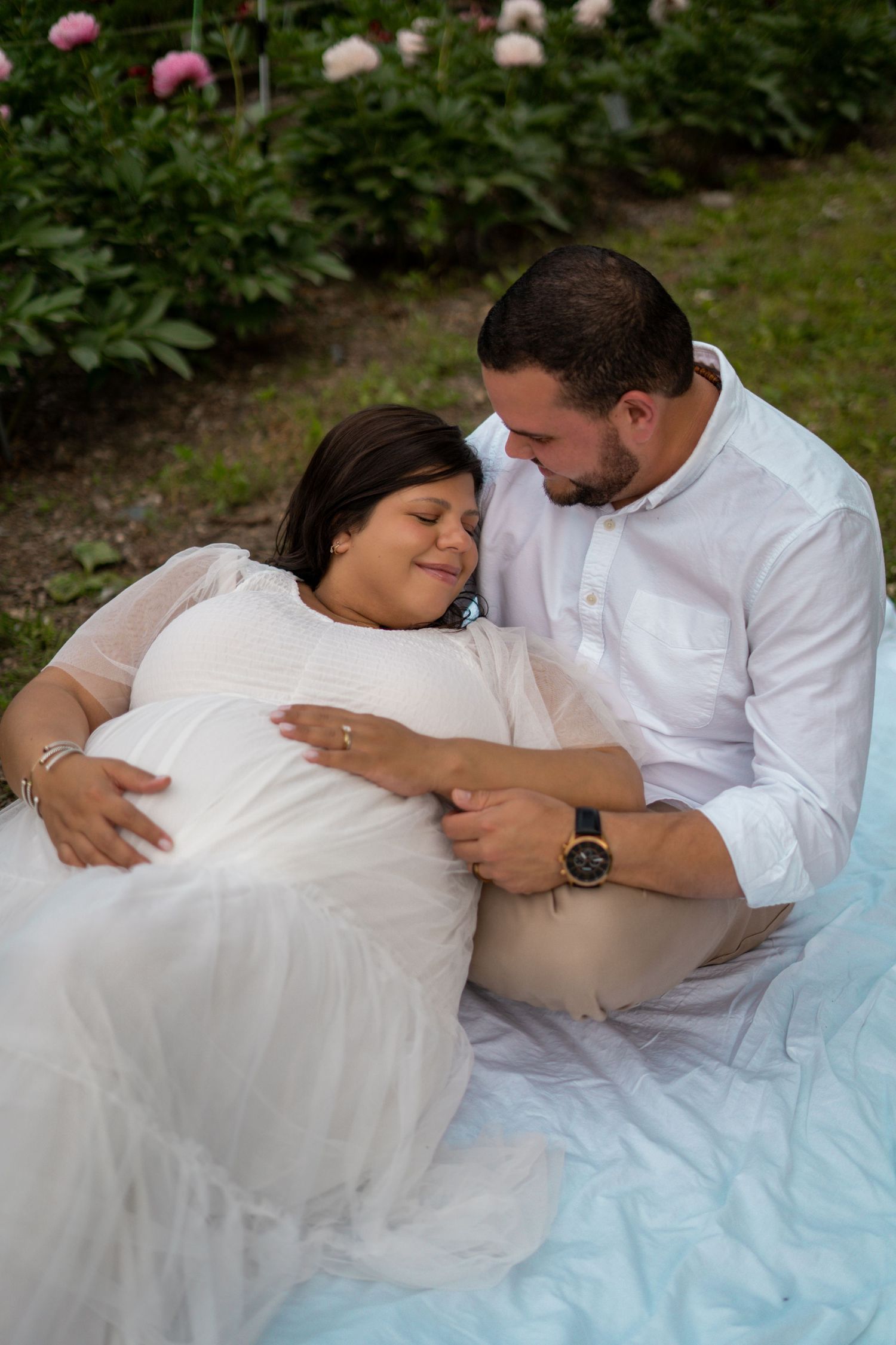 Couple sitting on a blanket in a garden, pregnant woman resting on partner's shoulder peacefully.