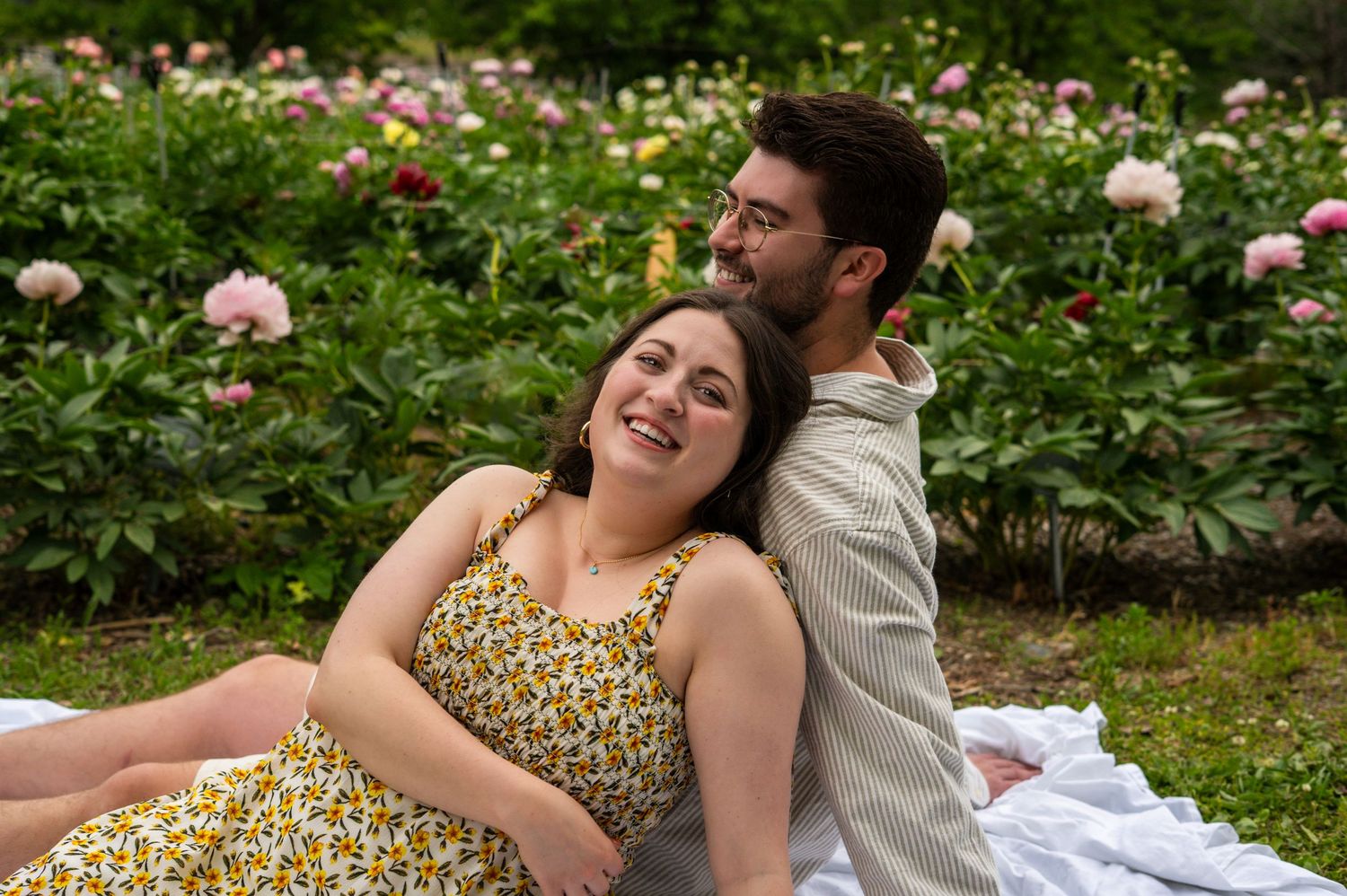 Happy couple sitting back-to-back on a white blanket surrounded by pink peony flowers in a lush garden.