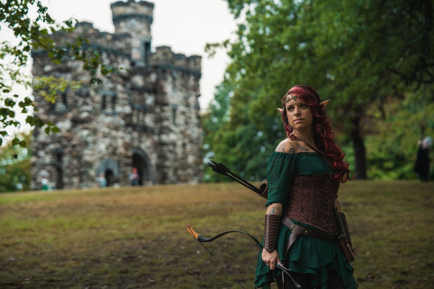 A woman in elf cosplay with red hair holds a bow near a stone castle, wearing a green dress and brown corset.