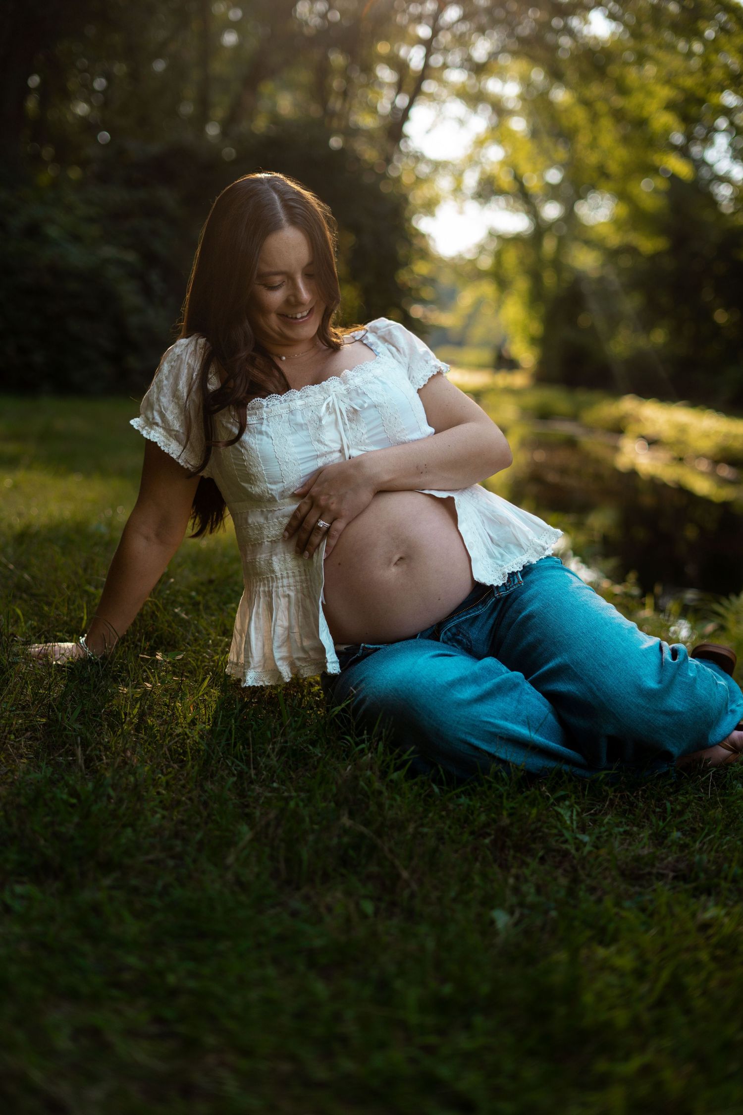 Pregnant woman in white top and jeans sitting on grass in golden sunlight touching her bare belly.