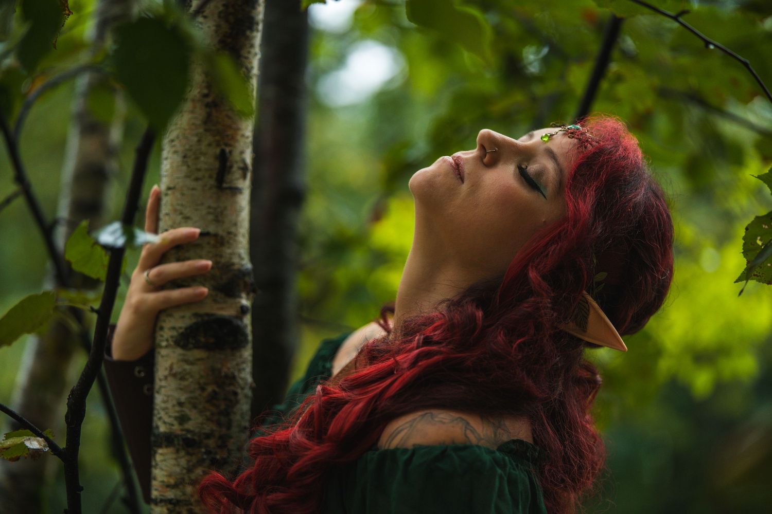A woman with long red hair and elf ears embraces a birch tree in a lush green forest wearing a medieval green dress.