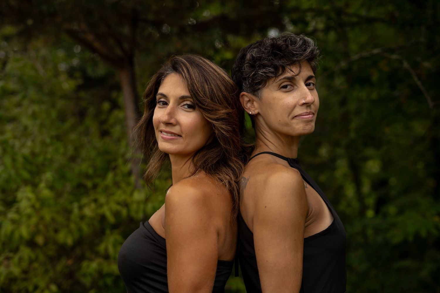 Two women in black tops standing back to back surrounded by lush green foliage.