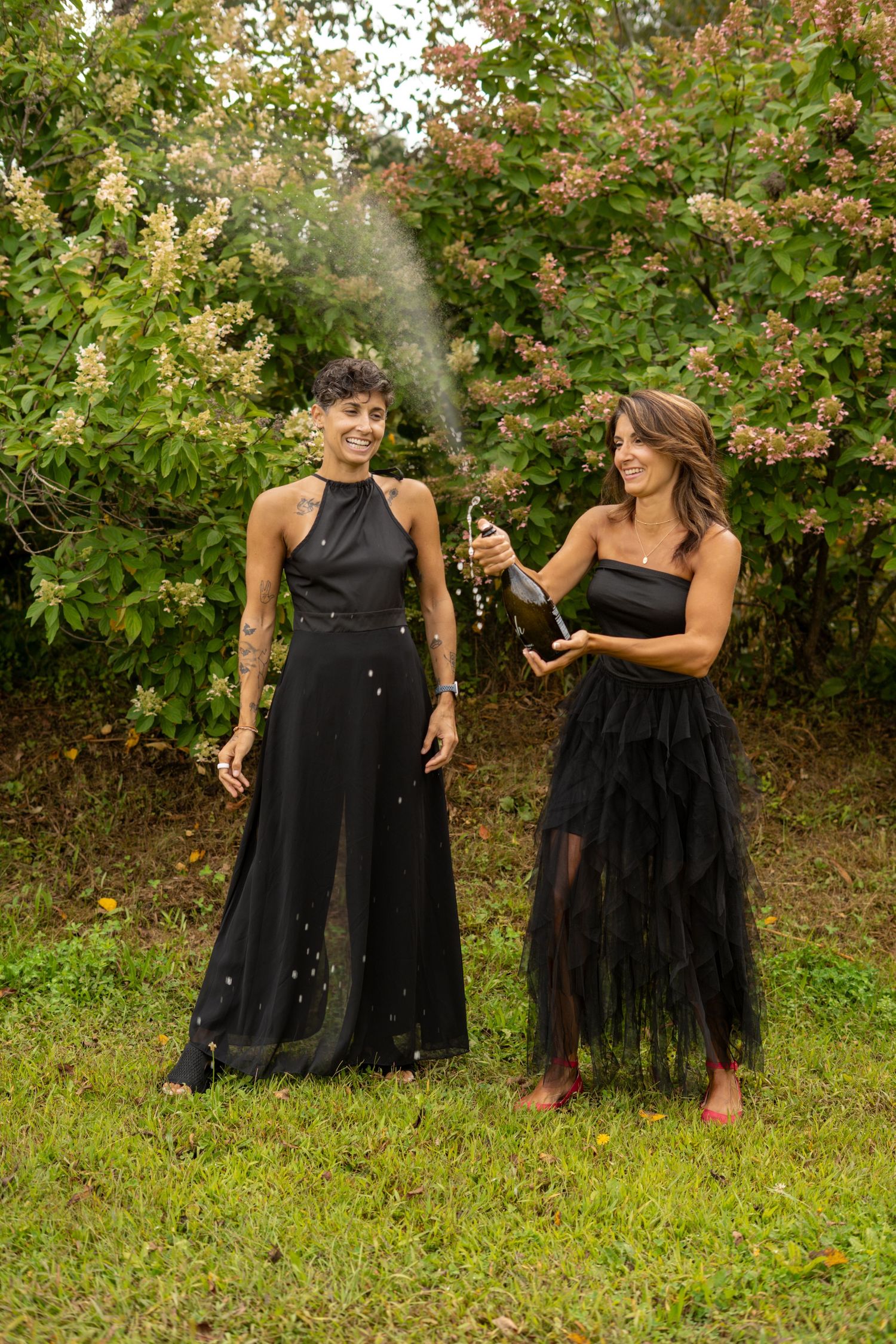 Two women in elegant black gowns popping a champagne bottle outdoors in a garden.