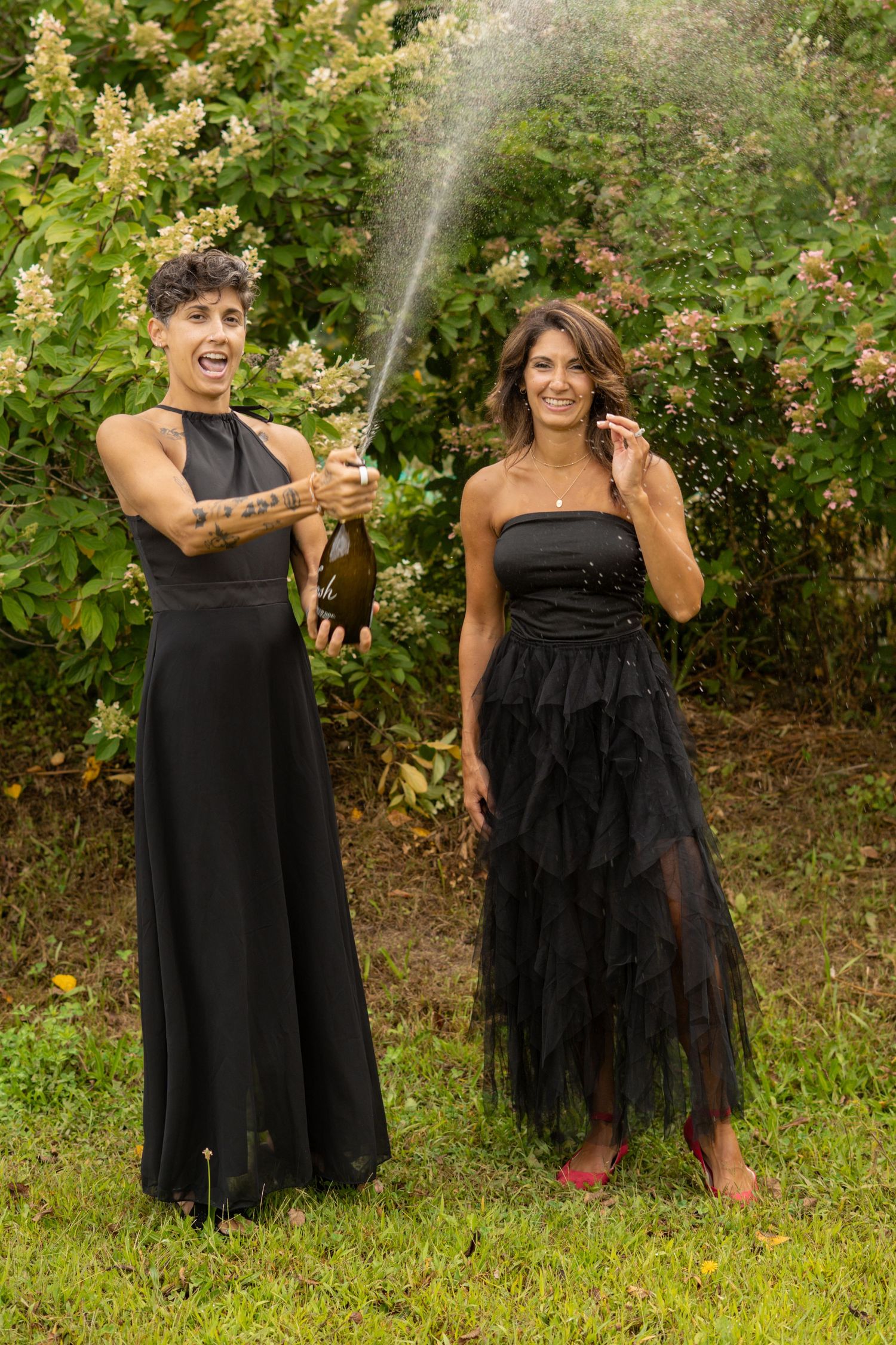Two women in black dresses celebrate outdoors, one spraying champagne near flowering bushes.