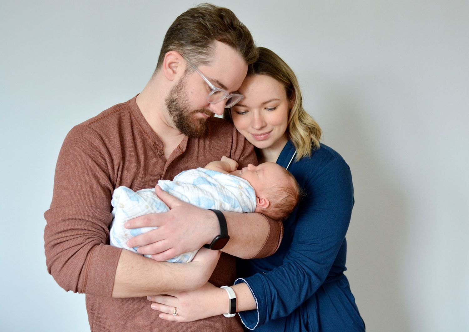 A new mom and dad pose with arms wrapped around each other and holding their newborn baby boy during a lifestyle photoshoot.