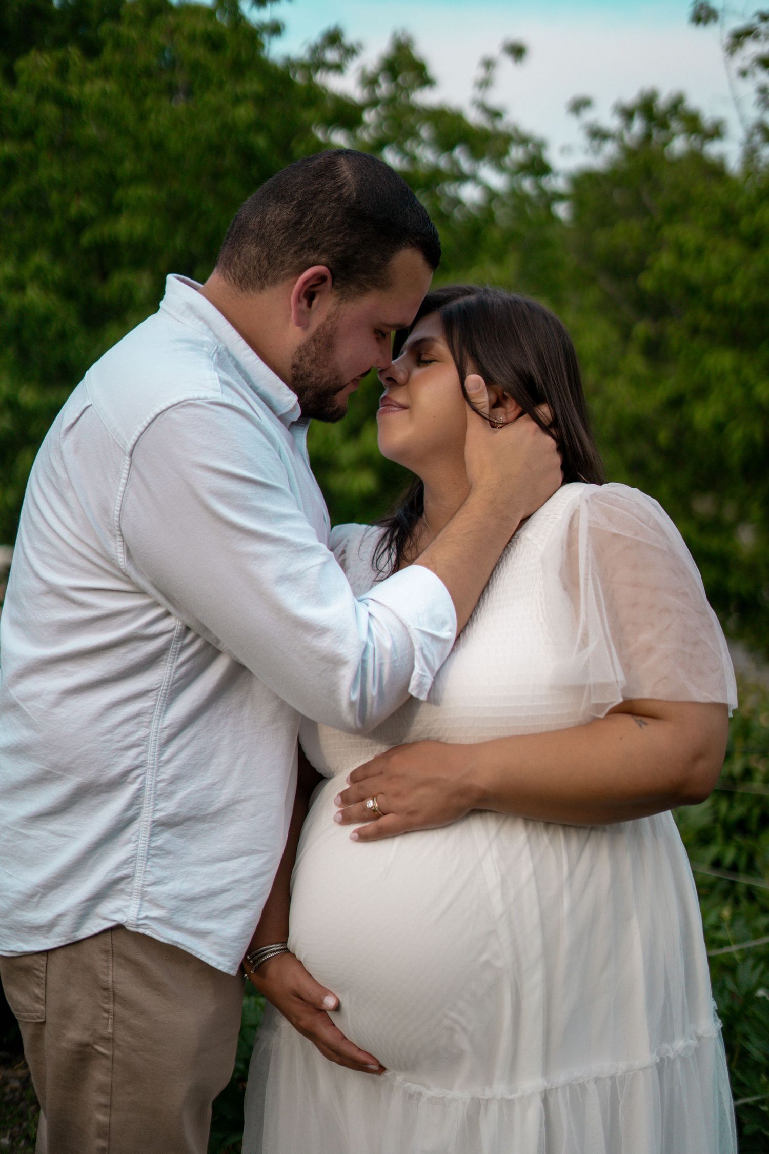 Couple shares tender moment outdoors, man gently holding pregnant womans face in white maternity dress.