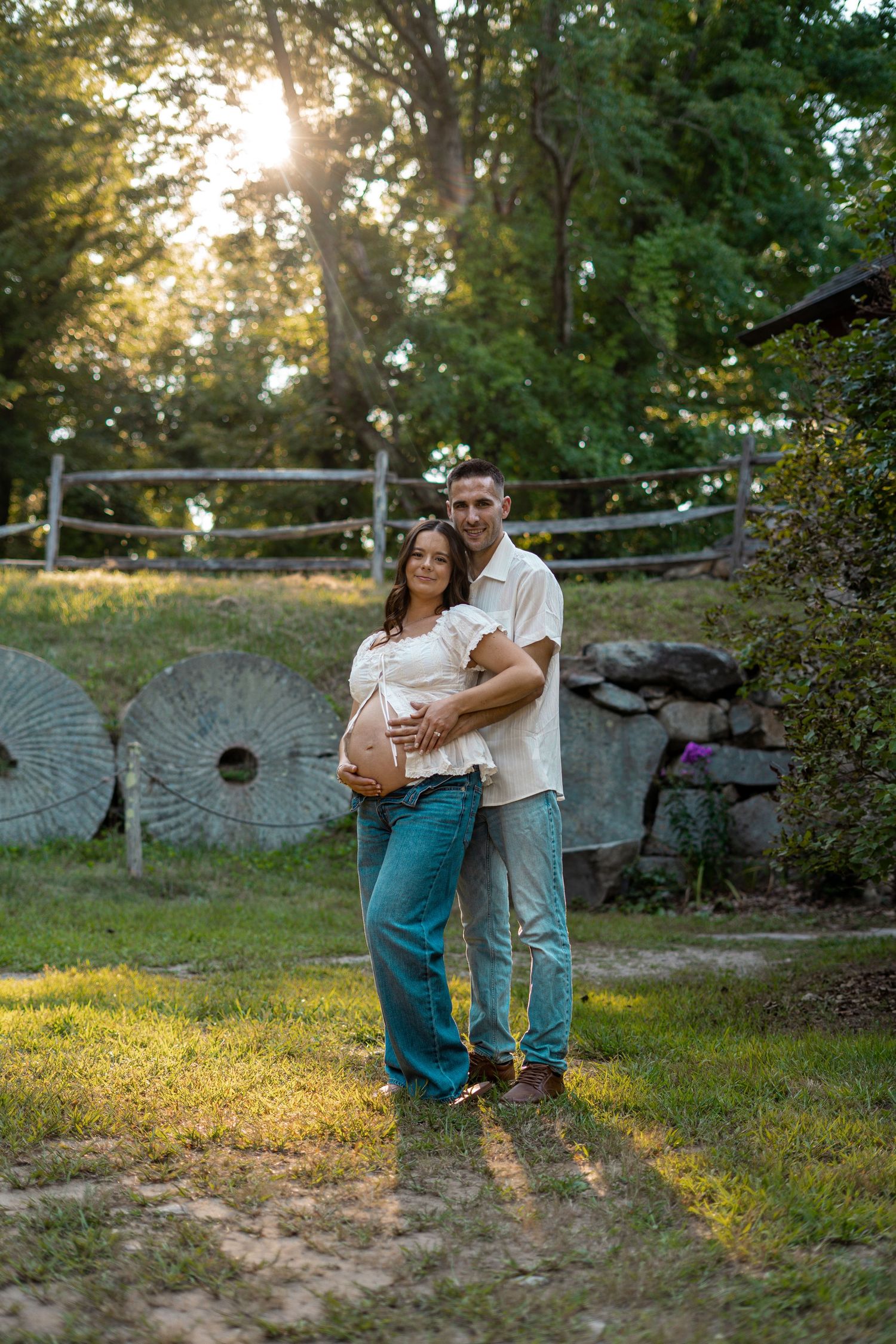Expecting couple embracing in a sunlit park maternity photo, the woman cradling her baby bump beside old stone mill wheels and a wooden fenc