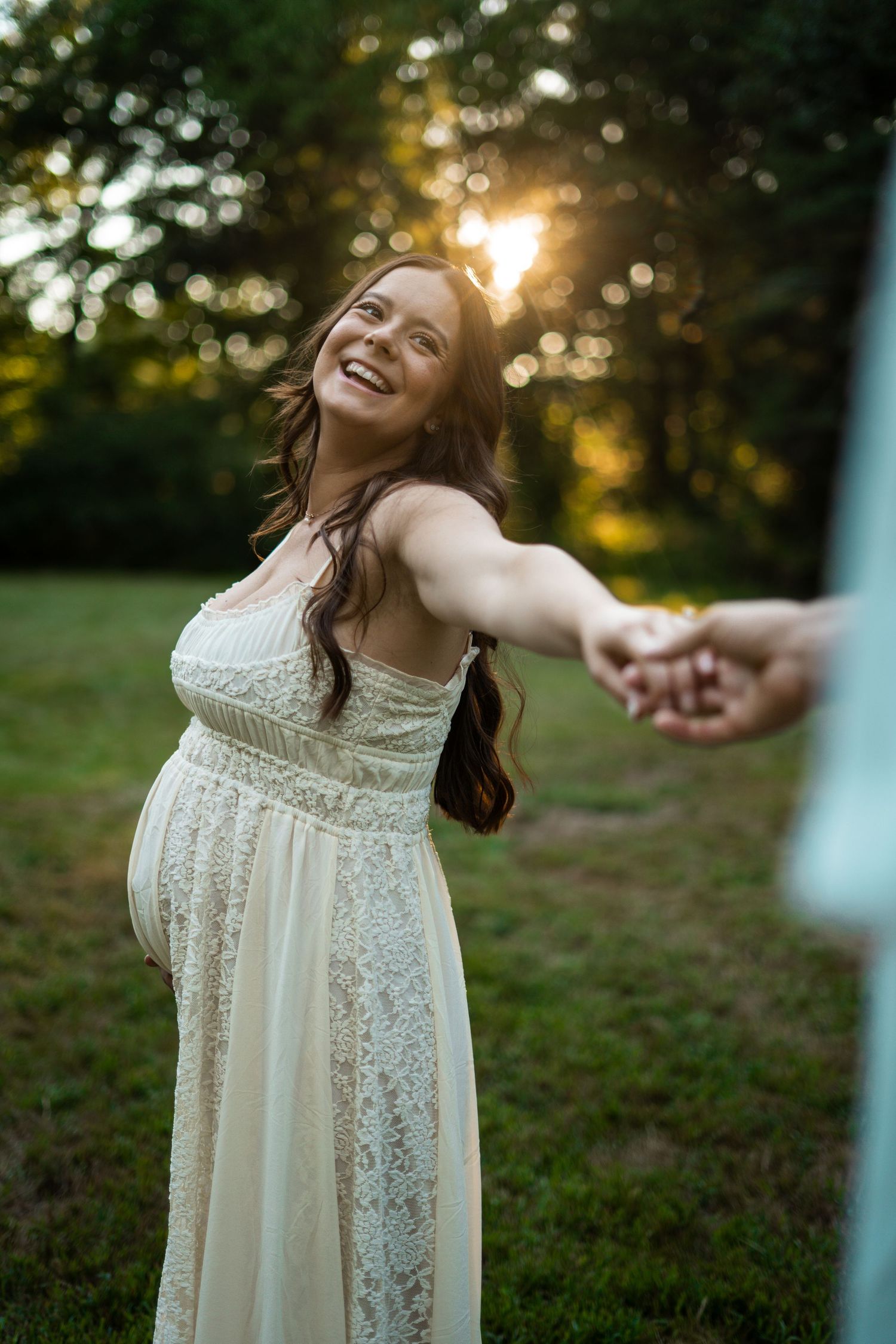 Smiling pregnant woman in white lace dress holding hands at golden sunset in a green park.