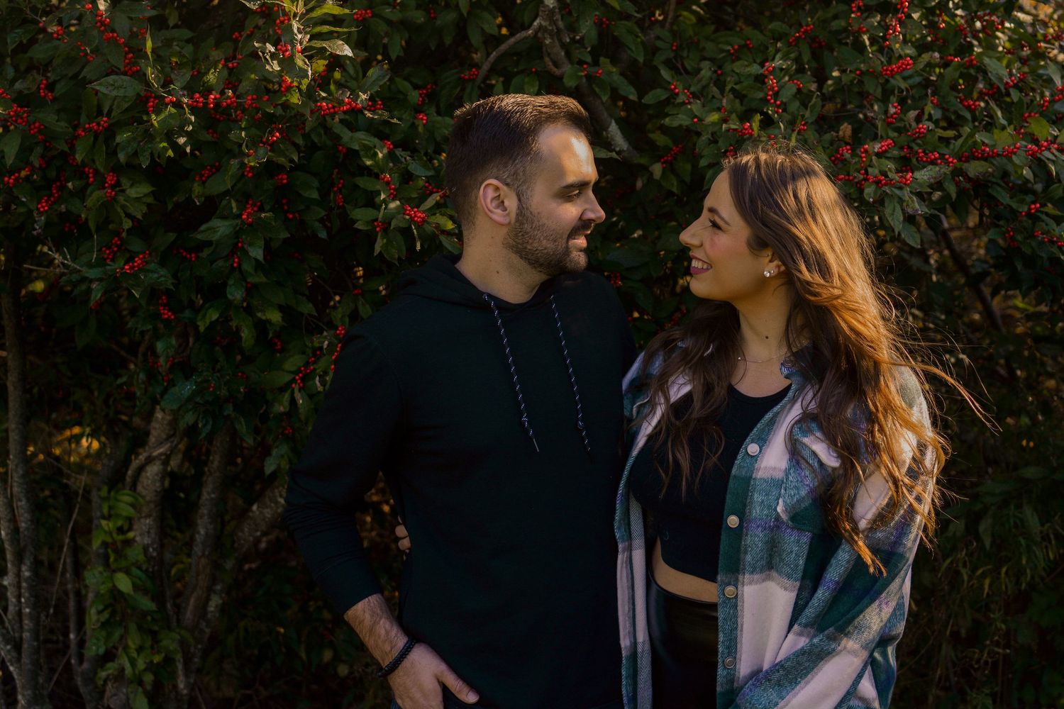 Smiling couple gazing at each other in front of lush red berry bushes at sunset.