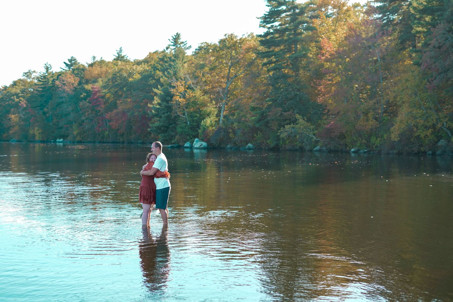 Couple standing together in a calm lake surrounded by vibrant autumn foliage.