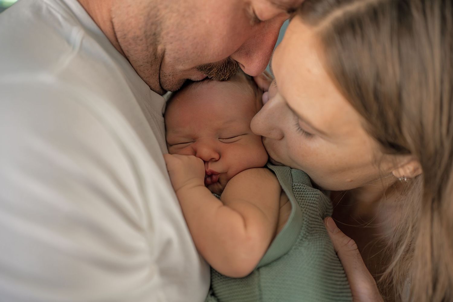 Loving parents gently kissing their sleeping newborn baby wrapped in a soft green swaddle.