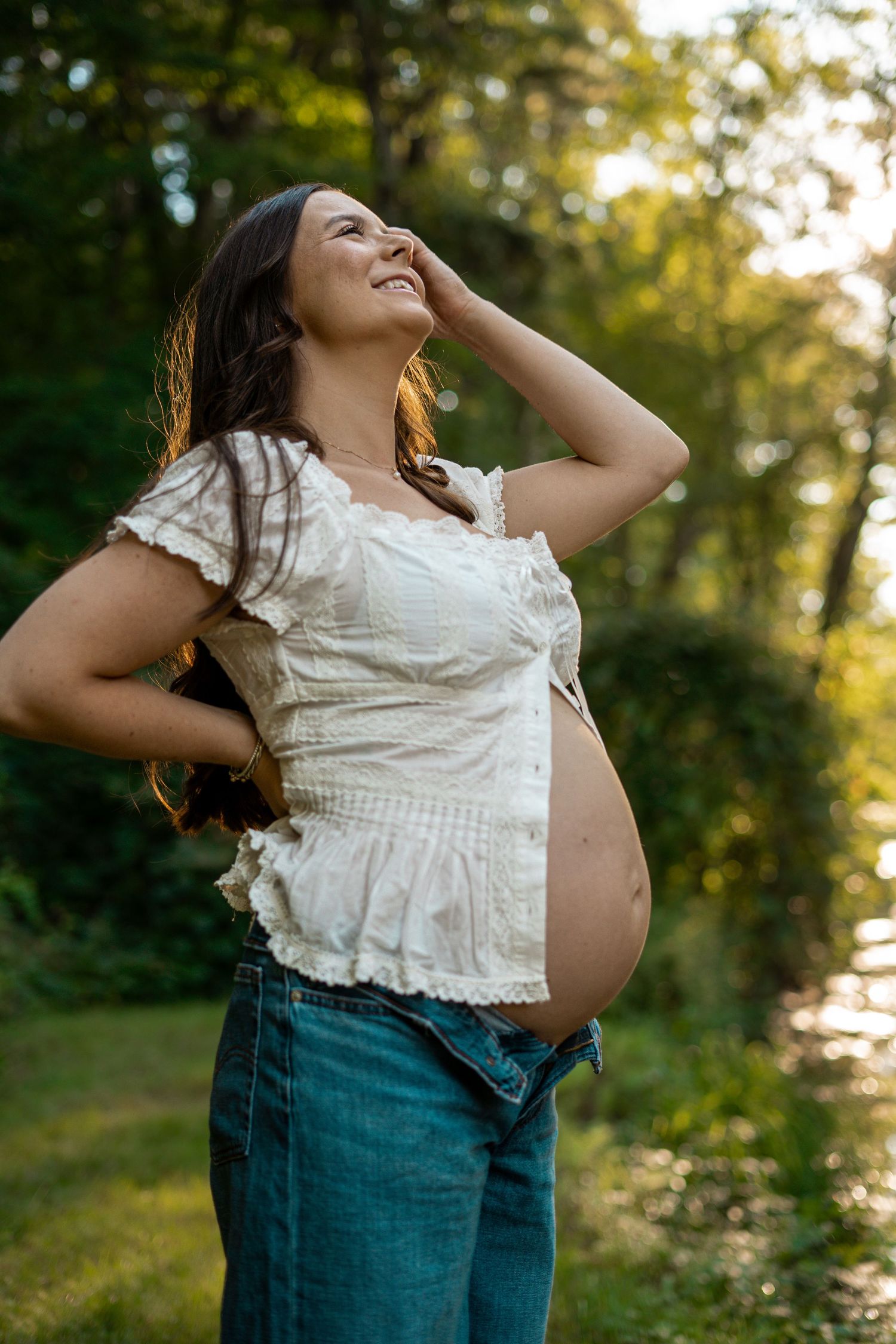 A smiling pregnant woman in a white crop top and jeans poses outdoors in golden sunlight.