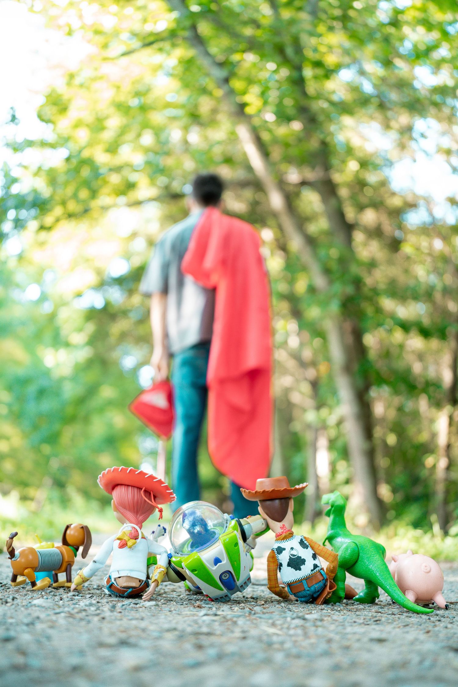 Toy Story figurines on gravel path with person in red cape walking away in woods.