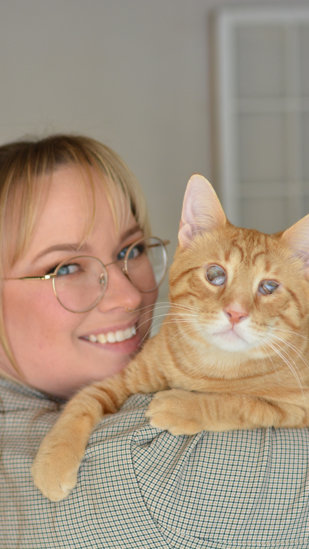 Smiling blonde woman with glasses holding an orange tabby cat close to her face indoors.