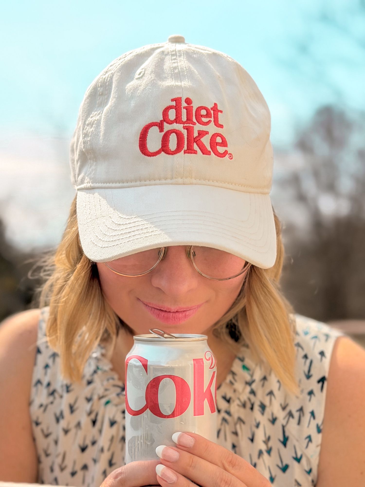 Blonde woman in a Diet Coke cap sipping from a Diet Coke can outdoors in sunlight.