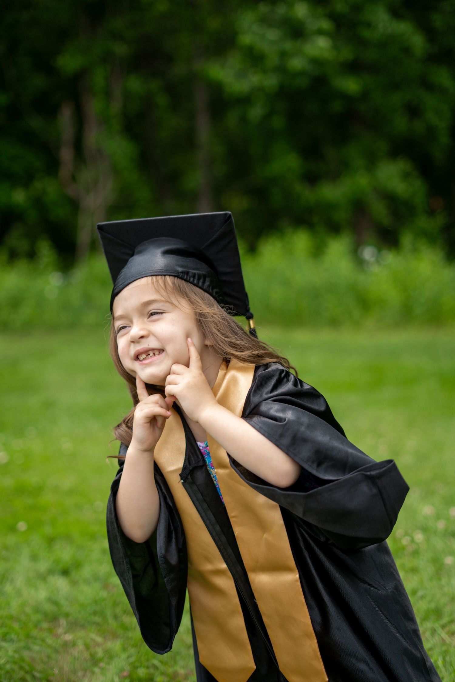 A joyful young girl in a black and gold graduation cap and gown poses outdoors in a green park.