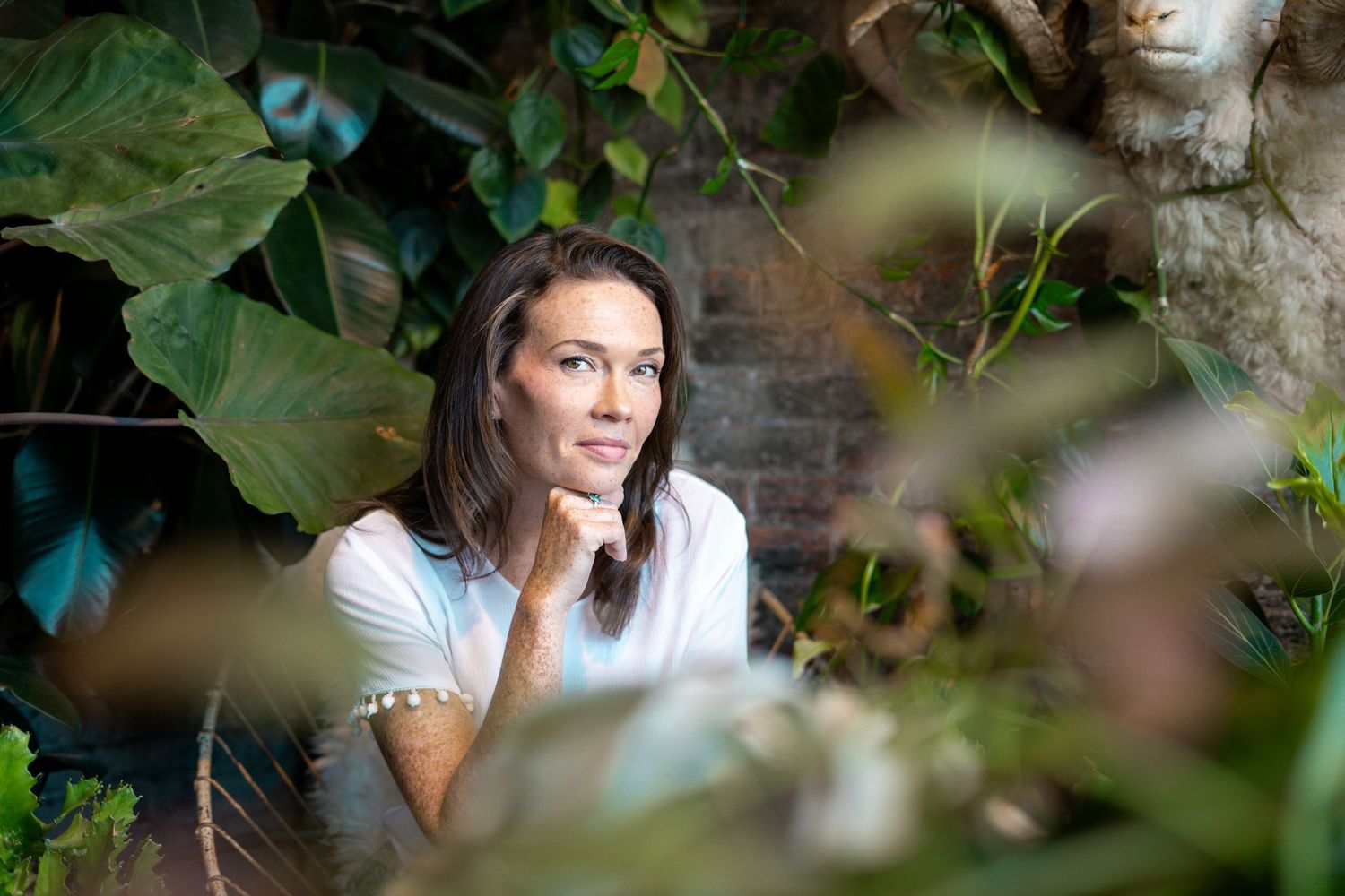 Thoughtful woman in white t-shirt sitting among tropical green plants in a garden setting.