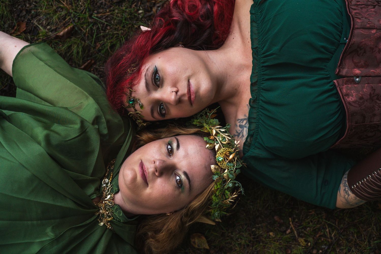 Two women in green dresses with floral crowns lying on grass in an enchanting forest-inspired photo shoot.