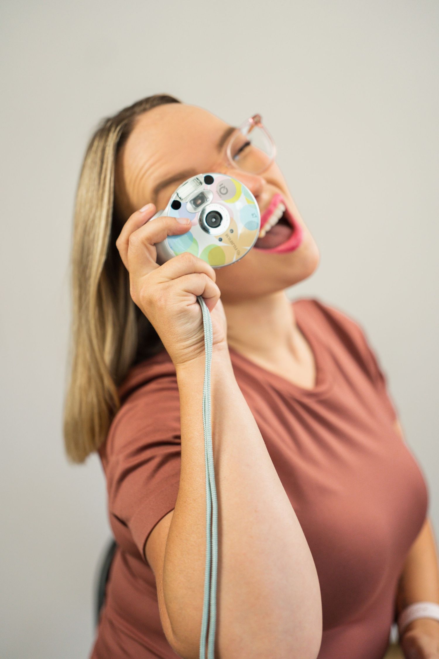 Laughing blonde woman holding a colorful retro camera up to her face against a neutral background.