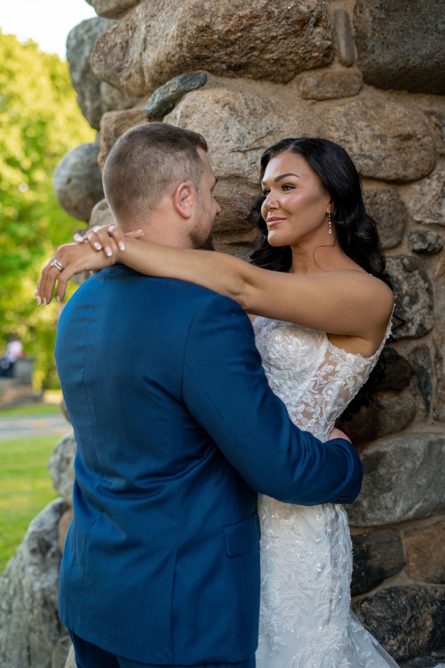 Bride in lace gown embracing groom against a stone wall, smiling warmly outdoors.